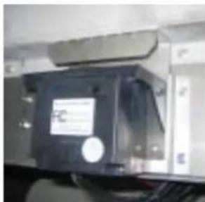 Close-up of a black electronic device with a label and 'FC' on its side, mounted on a metal frame (no readable text or symbols beyond the label)