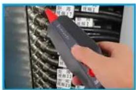 Close-up of a hand holding a pen tip next to a rack of cables with visible labels and red connectors