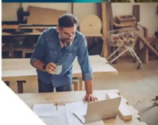 Man in blue shirt standing at a desk with a laptop, working in a workshop with wooden furniture and no visible text or symbols.
