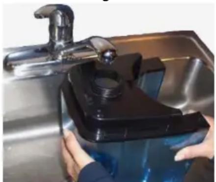 Person washing a blue plastic tank into a stainless steel kitchen sink (no text or symbols visible)