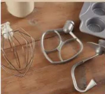 Close-up of kitchen utensils including a wire mixer, metal mixing bowl, and stand mixer on a wooden surface (no text or symbols visible)