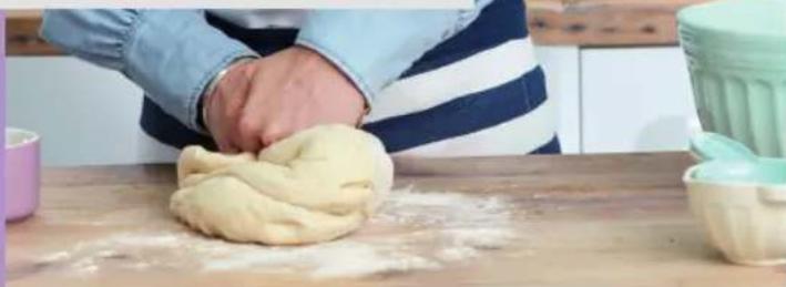 Person kneading dough on a wooden table with flour and a cup nearby (no text or symbols visible)