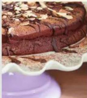 Close-up of a chocolate cake with powdered sugar topping, served on a white fluted stand (no text or symbols visible)