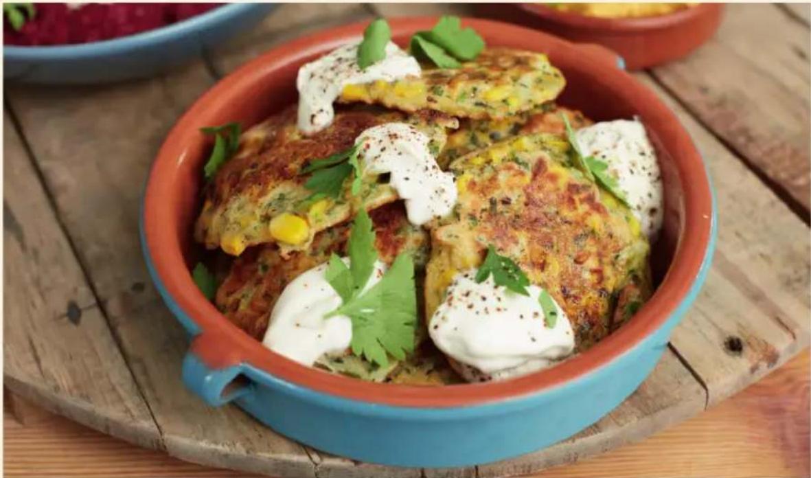 Close-up of a colorful ceramic dish with baked potatoes, corn, and herbs in a red ceramic bowl, placed on a wooden table (no text or symbols visible)