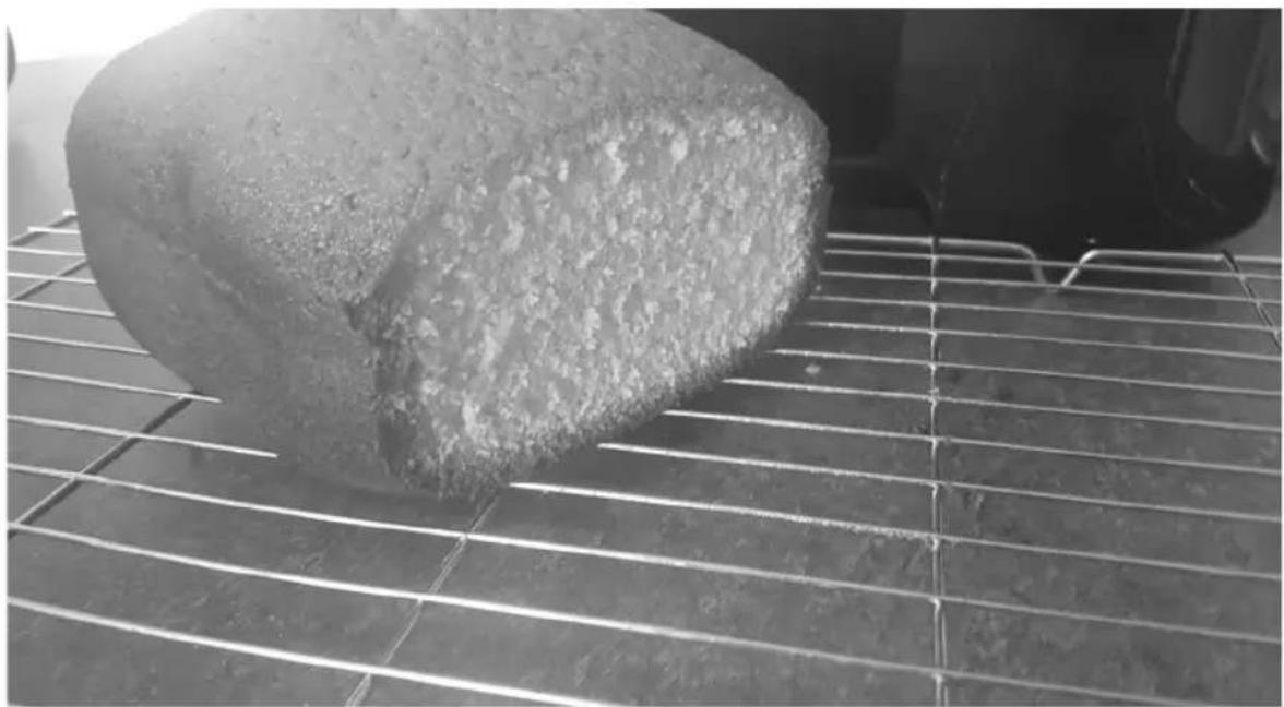 Close-up of a large, irregularly shaped food item on a cooling rack, with a blurred background showing a dark object (no text or symbols visible)