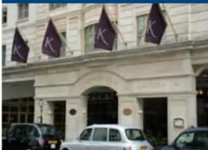 Street photo showing a building facade with multiple purple flags bearing the letters 'K' and visible signage, alongside parked cars.