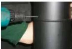 Close-up of a hand using a tool to apply material, with a dark cylindrical container and green cloth partially visible (no text or symbols)