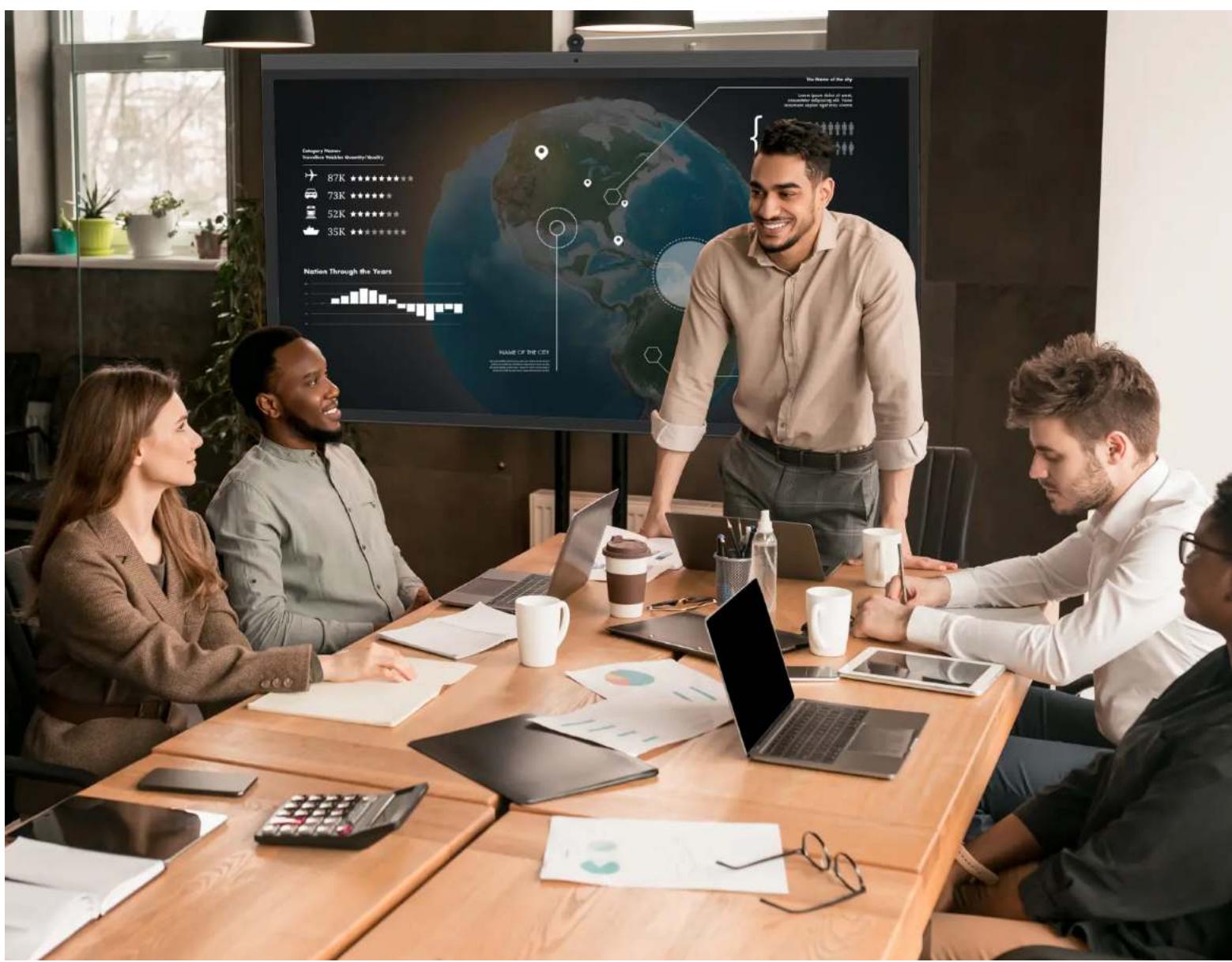Conference room scene with a presenter pointing at a large digital dashboard displaying cityscape metrics and data, surrounded by business team members.