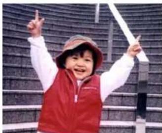 A smiling child in a red vest and hat, raising one finger with both index fingers, against a textured wall background (no text or symbols visible).