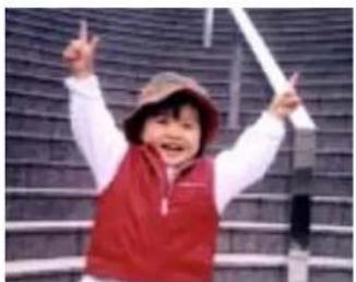 Child wearing red vest and hat, smiling with raised arms in front of a textured wall (no visible text or symbols)