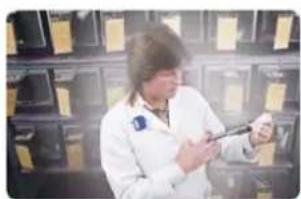 A person in a white lab coat holding a small object, standing in front of shelves with framed certificates (no visible text or symbols)
