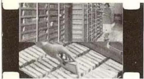Black-and-white photo of a person handling a large object in a storage room with shelves and pallets (no visible text or symbols)
