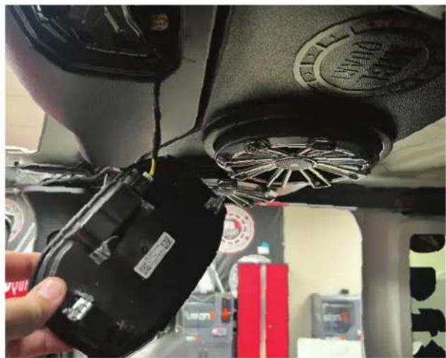 Close-up of a hand holding a black electronic device with visible wiring and a circular component, in a vehicle interior (no text or symbols)