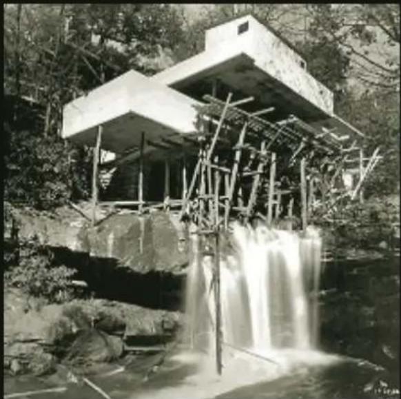 Black-and-white photo of a modern concrete structure with waterfalls and wooden scaffolding, surrounded by trees (no visible text or symbols)