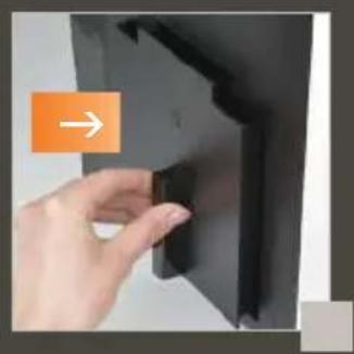 Hand holding a small dark object next to a black panel, with an orange arrow pointing to it (no text or symbols visible)