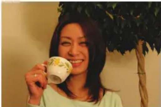 Woman smiling while holding a decorative teacup, with leafy tree in background (no text or symbols visible)