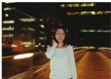 A person posing on a wooden bridge at night with illuminated buildings in the background (no visible text or symbols)
