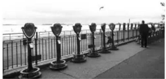 Black-and-white photo of a waterfront promenade with multiple public payee barriers and a person standing on the railing, no visible text or symbols.