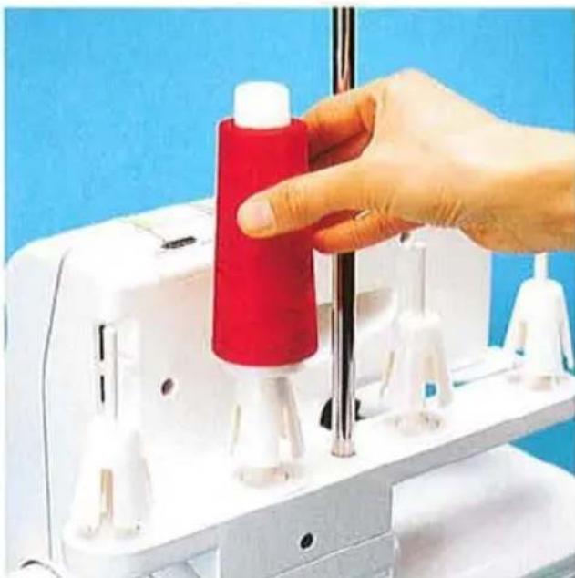 Main actionnant une machine à coudre avec une bobine de fil rouge, sur un fond bleu (aucun texte ou symbole visible)
