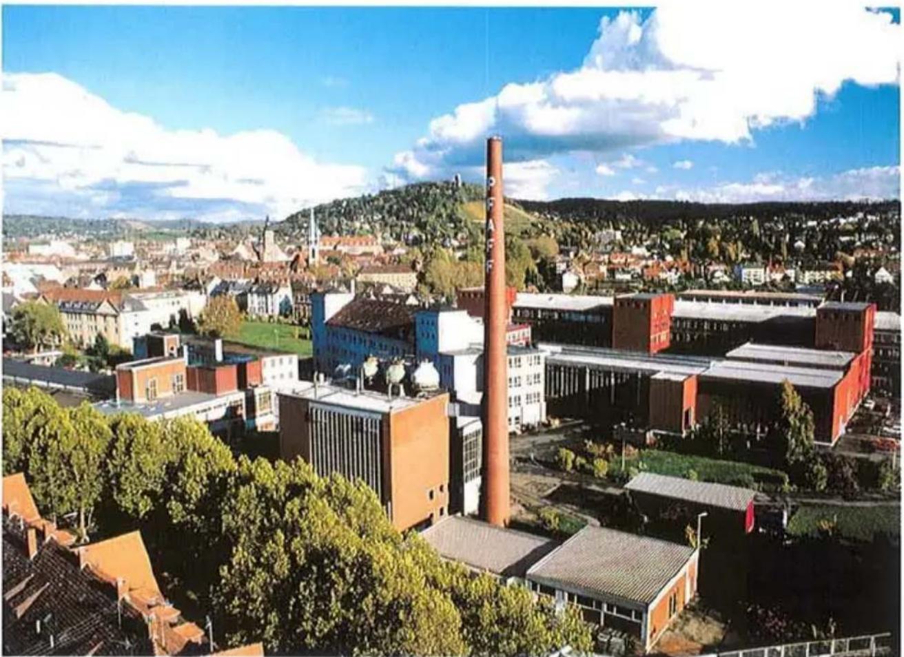 Vue aérienne d'un complexe industriel moderne avec une cheminée proéminente et de la verdure environnante sous un ciel bleu (aucun texte ou symbole visible)