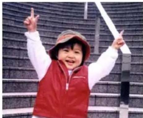 A smiling child in a red vest and hat, raising one finger with both hands, standing in front of a patterned stone structure (no text visible).