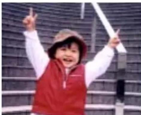 A smiling child in a red vest and hat, raising one hand with both fingers, against a dark tiled wall background (no text or symbols visible).
