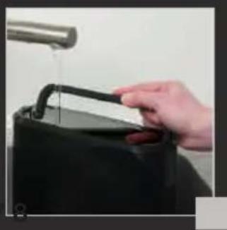 Hand pouring liquid from a black container into a stainless steel faucet (no text or symbols visible)