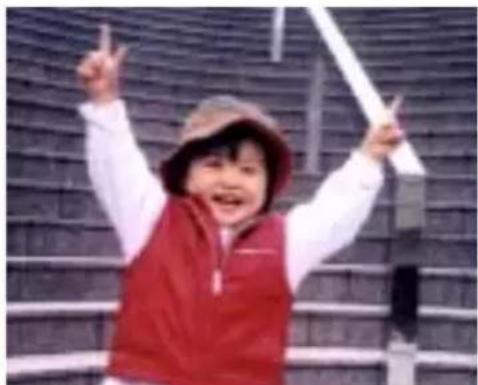 Child in red vest and hat celebrating with raised arms, standing in front of a brick wall (no text visible)