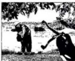 Two people outdoors near a water body, one holding a tool, under trees (no visible text or symbols)