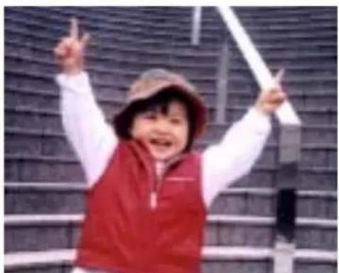 A smiling child in a red vest and hat, raising one hand with both fingers, against a dark tiled wall background (no text or symbols visible).