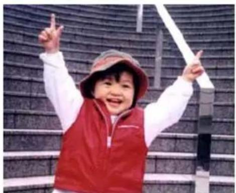 A smiling child in a red vest and hat, posing with raised fists in front of a patterned metal staircase (no text visible)