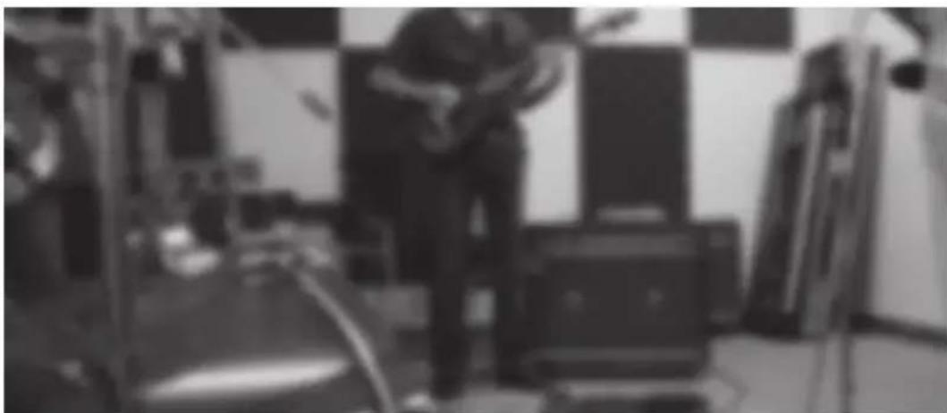 Black-and-white photo of a person playing a guitar in a dimly lit indoor space with equipment and no visible text or symbols.