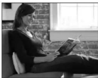 Black-and-white photo of a woman sitting on a couch reading a book, with brick wall background (no visible text or symbols)