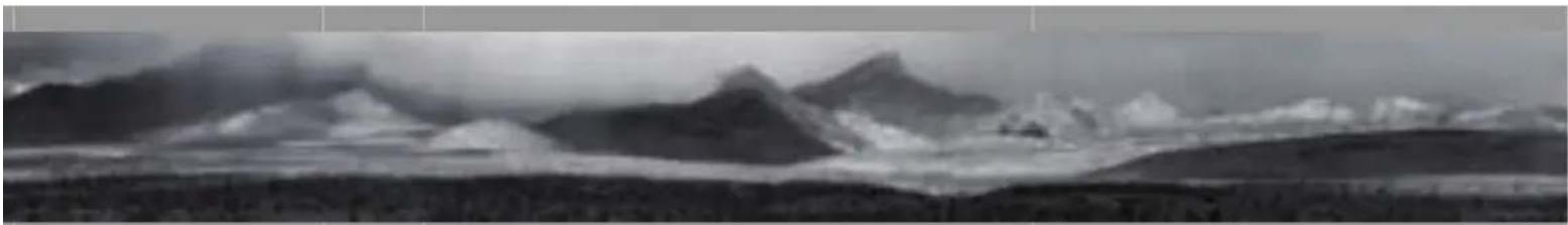 Panoramic black-and-white landscape photo showing snow-capped mountains rising above a valley with forested slopes below (no text or symbols)