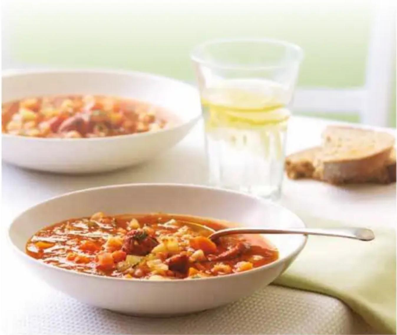 Bowl of tomato sauce with a spoon, bread slices, and a glass of juice in the background (no text or symbols visible)