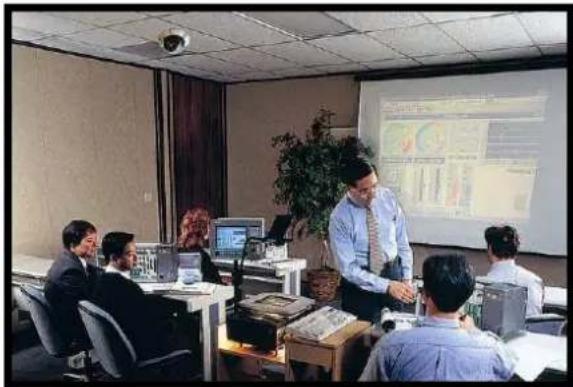 Interior view of a modern office with people working at desks and a presenter at the front, projected screen visible (no readable text or symbols)