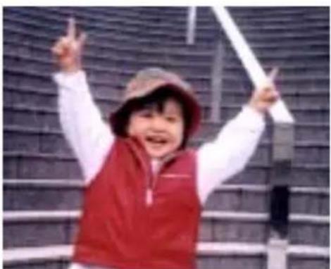 Child in red vest and hat raising fists in front of a brick wall (no visible text or symbols)