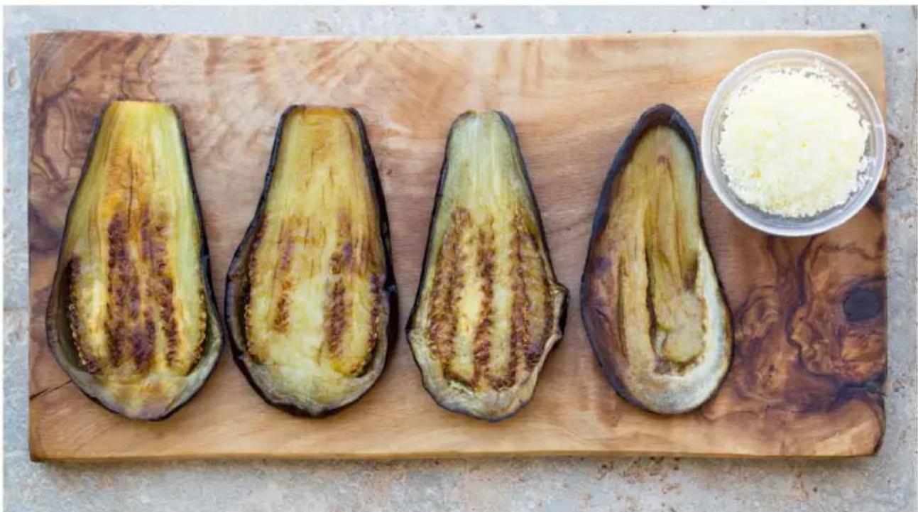 Four eggplant halves on a wooden cutting board, with a small bowl of white powder in the background (no text or symbols visible)