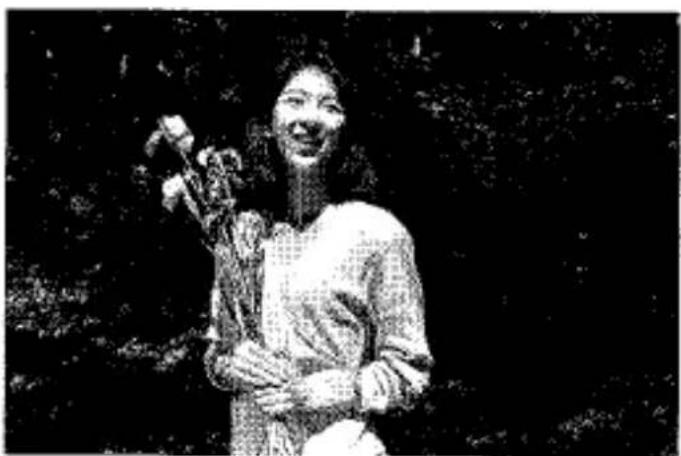 Photo en noir et blanc d'une personne souriante tenant un objet, à l'extérieur avec un feuillage flou en arrière-plan (aucun texte ou symbole visible)