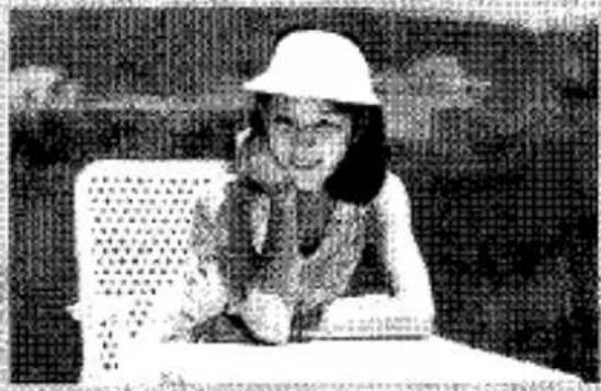 Photo en noir et blanc d'une fille souriante portant un chapeau et tenant un objet, assise à une table avec un fond à motifs (aucun texte ou symbole visible)