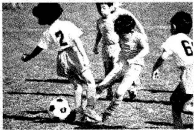 Photo en noir et blanc d'un match de football avec des joueurs sur le terrain et un ballon au sol (aucun texte ni symbole visible)