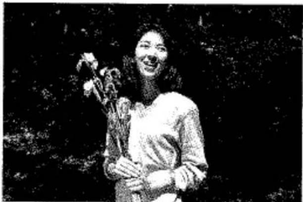 Photographie en noir et blanc d'une femme souriante tenant des fleurs, à l'extérieur avec un arrière-plan de feuillage flou (aucun texte ou symbole visible)