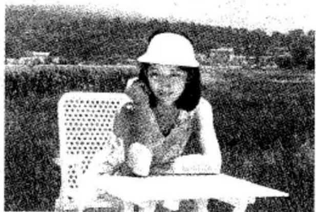 Photo en noir et blanc d'une personne assise à une table à l'extérieur, portant un chapeau et une chemise à col, avec des arbres et des bâtiments au loin en arrière-plan (aucun texte ni symbole visible)