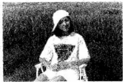 Photographie en noir et blanc d'une personne assise sur une chaise, portant une chemise blanche avec un t-shirt à motifs (aucun texte ou symbole visible)