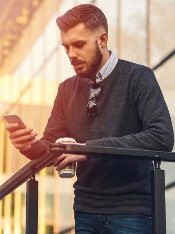 Man with beard using smartphone on balcony railing, holding coffee cup (no visible text or symbols)