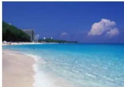 Tropical beach scene with turquoise water, sandy shore, and distant buildings under a clear blue sky (no text or symbols visible)