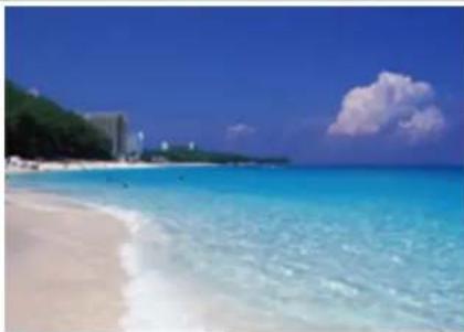 Coastal beach scene with turquoise water, sandy shore, and distant buildings under a clear blue sky (no text or symbols visible)