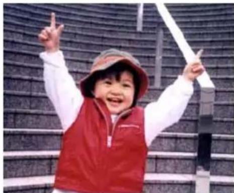 A smiling child in a red vest and hat, raising one finger with both hands, against a blurred stadium or building facade (no visible text or symbols).