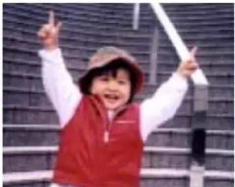 Child wearing red vest and hat, smiling with raised arms in front of a brick wall (no visible text or symbols)