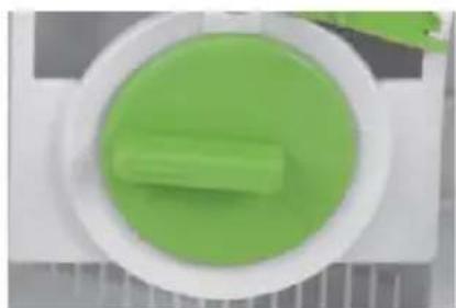Top-down view of a green cylindrical object inside a white circular container, placed on a metal plate (no text or symbols visible)